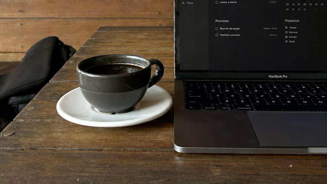 Una taza de café junto a un portátil abierto sobre una mesa de madera.